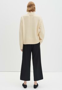 Beiger Strickpullover mit hohem Kragen, geripptem Saum und heruntergesetzten Schulternähten, kombiniert mit schwarzen, weit geschnittenen Hosen und schwarzen Ballerinas.