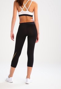 Czarne legginsy capri z złotą linią akcentową, zestawione z białym sportowym biustonoszem z krzyżującymi się paskami na plecach. Gładka, elastyczna tkanina.