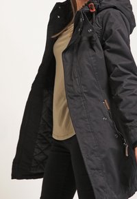 Sort, lang parka med hætte, trykknaplukning og en sidelomme med lynlås. Den har en quiltet foring og en justerbar snor i taljen.