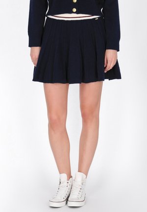 Modelo con una minifalda plisada azul marino y una chaqueta de punto a juego con botones dorados, combinadas con zapatillas blancas de caña alta.