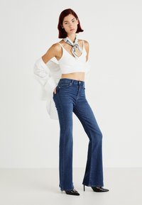 Μπλε φαρδειά τζιν με ψηλή μέση, φτιαγμένα από τζιν ύφασμα. Συνδυάζονται με λευκή μπλούζα crop και ένα μοτίβο μαντήλι γύρω από τον λαιμό.