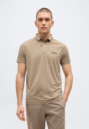 Jeune homme portant un polo BOSS beige et un pantalon assorti, debout avec une main dans la poche, sur un fond clair uni.