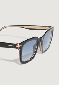 Gafas de sol de plástico negro con una montura gruesa, lentes azules degradados, acentos dorados y rayas rojas y blancas en las patillas.