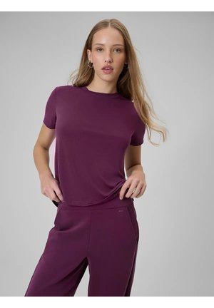 Młoda kobieta w dopasowanym fioletowym krótkim topie i spodniach, pozująca z rękami przy talii na jednolitym szarym tle.