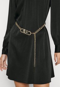 Czarna sukienka z długimi rękawami, zestawiona ze złotym paskiem łańcuchowym z klamrą w kształcie logo "MK" oraz zwisającym detalem w postaci łańcucha.