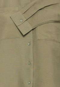 Primo piano di una camicia beige a maniche lunghe con polsini abbottonati, bottoni frontali e due tasche sul petto con cuciture visibili.