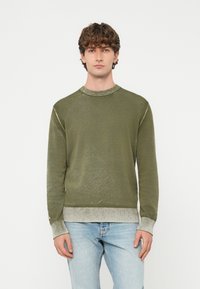 Jauns vīrietis ar cirtainiem matiem, valkā olīvzaļu džemperi ar gaiši pelēkām aprocēm un apakšu, nēsā gaiši zilas džinsas, skatās taisni priekšā.