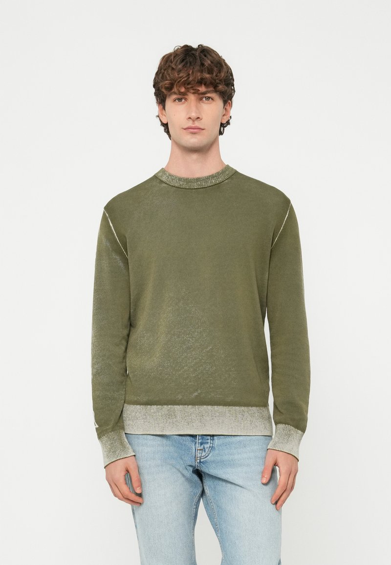 Jauns vīrietis ar cirtainiem matiem, valkā olīvzaļu džemperi ar gaiši pelēkām aprocēm un apakšu, nēs�ā gaiši zilas džinsas, skatās taisni priekšā.