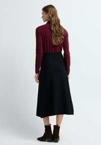 Bordeaux lange mouwen top gecombineerd met een zwarte, knielange rok. Het model draagt donkerbruine suede enkellaarzen met een blokhak. Soepele texturen overal.