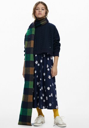 Maglione a trecce blu navy, sciarpa a quadri con tonalità di verde e marrone, vestito scuro a fantasia floreale, calzini gialli e sneakers bianche.