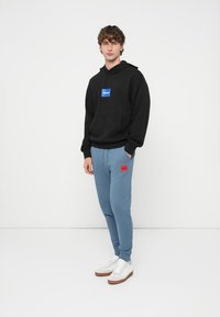 Mladý muž stojaci oblečený v čiernej mikine s kapucňou a modrým logom, modrých teplákoch s červeným logom a bielych teniskách na bielom pozadí.