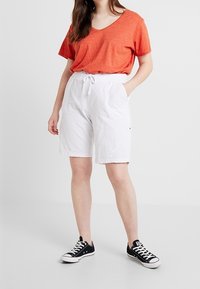 T-shirt orange à col en V, shorts blancs à taille élastique avec poches, et baskets noires avec semelles blanches. Tenue décontractée d'été.