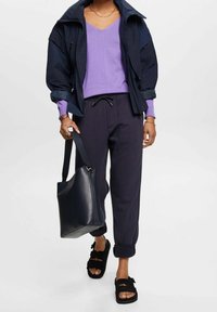 Granatowa kurtka wiatrówka, jasnofioletowy sweter z dekoltem w serek, ciemne dresy z mankietami na kostkach, czarna torba na ramię i czarne sandały z klamrami.