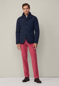 Giacca trapuntata blu navy con colletto alto, chiusure a scatto e due tasche frontali, abbinata a pantaloni rossi e stivali neri. Sfondo semplice.