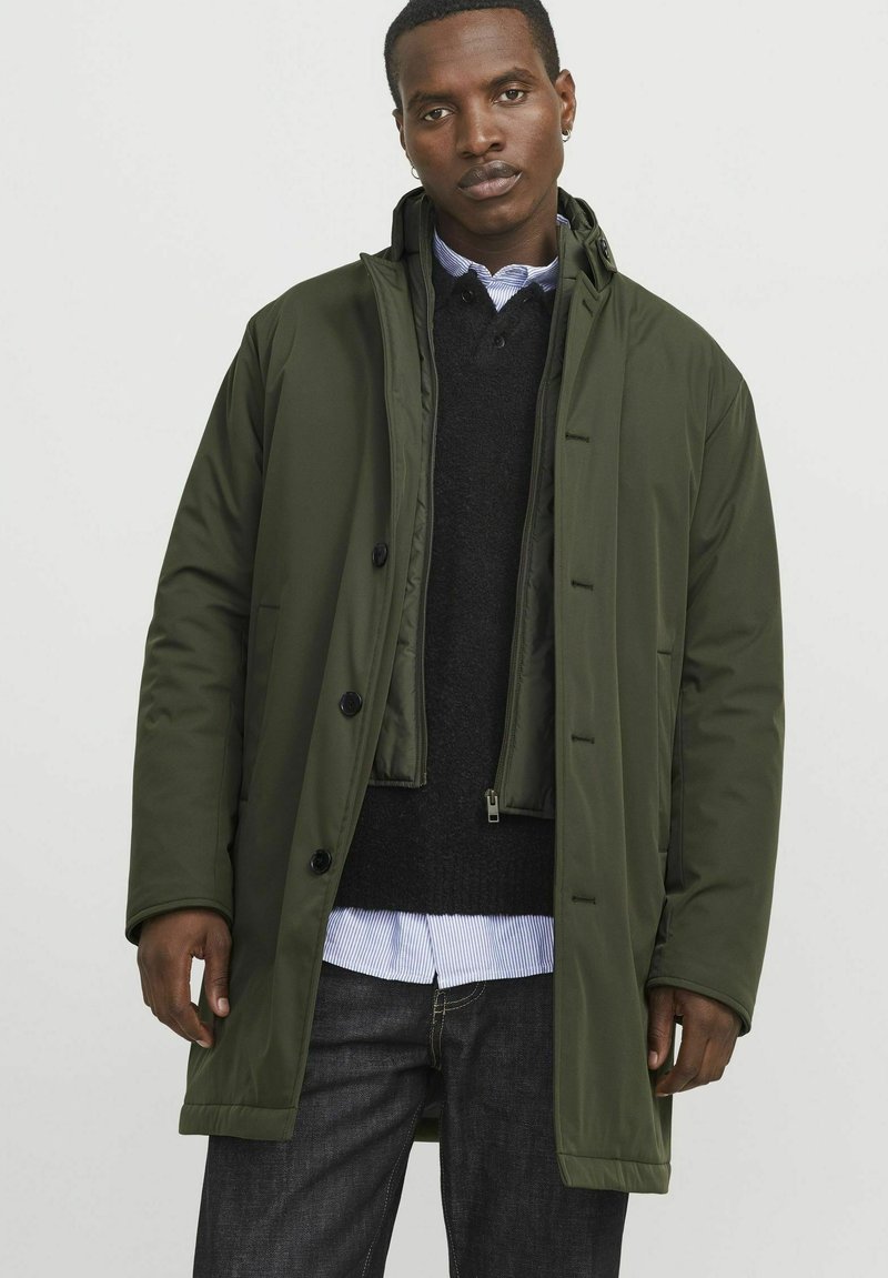 Giacca isolante verde oliva con chiusura a bottoni, cappuccio removibile e tasche laterali. Indossata sopra un maglione nero e una camicia a righe blu.