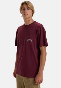 Bordeaux kortærmet t-shirt med lomme, der har hvid broderet tekst på brystet, lavet af blødt bomuldsstof.