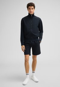 Marineblaue Sportjacke mit hohem Kragen und Reißverschluss, kombiniert mit passenden Shorts. Das Material hat eine strukturierte Optik. Weiße Slipper.