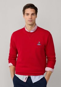 Felpa rossa con collo a giro e maniche lunghe. Presenta un logo ricamato blu e bianco. Indossata sopra una camicia a righe blu e bianca.