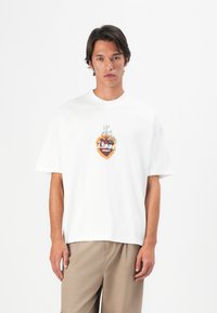 Jonge man in een wit oversized T-shirt met een vlammenhart en bloemgraphic, gecombineerd met beige wijde broekspijpen.