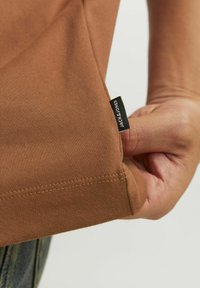 Hnědé bavlněné tričko s měkkou texturou, s černou etiketou "JACK & JONES" na lemu, ukazující detaily šití.
