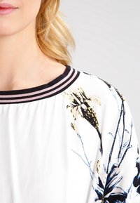 Vitt plagg med blommönster i blått och gult. Har en svart och rosa randig halsringning. Slät textur, lös passform.