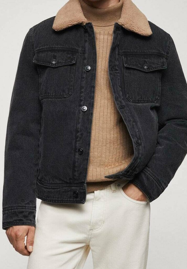 Homme portant une veste en jean noire avec un col en shearling beige sur un pull côtelé beige et un pantalon blanc, main dans la poche.