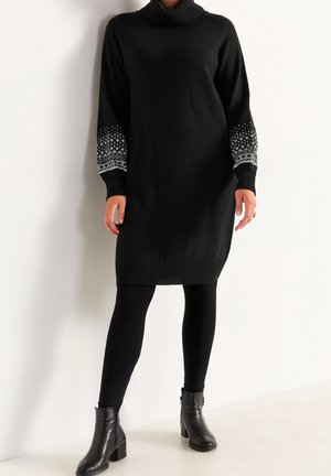 Kvinde iført en sort strikket kjole med hvide mønstrede manchetter, sorte leggings og sorte ankelstøvler, der står op ad en hvid væg.