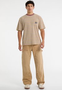 Gestreiftes kurzärmliges T-Shirt in Beige, mit lila und senfgelben Linien; kombiniert mit lockeren khakifarbenen Hosen mit seitlichen Taschen und Knopfakzenten.
