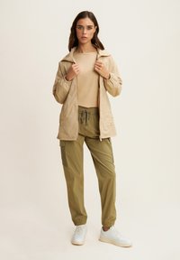 Chaqueta ligera beige con cierre de cremallera y puños elásticos, combinada con pantalones verde oliva con bolsillos laterales y elásticos en los tobillos.