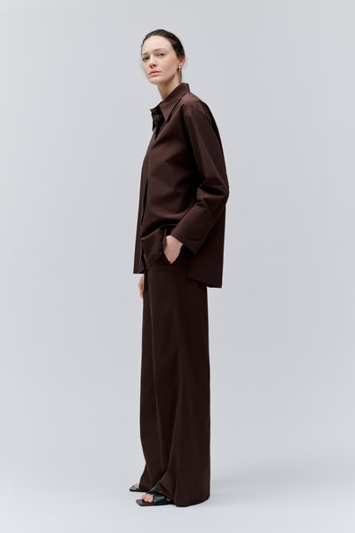 Femme debout dans une chemise oversize marron foncé et un pantalon à jambes larges, une main dans la poche, sur un fond gris clair.