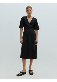 Zwarte midi-jurk met een diepe V-hals, korte ruffles mouwen, een knoopsluiting aan de voorkant en een zijsplit, gemaakt van lichte stof. Platte zwarte sandalen.