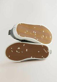 Een paar grijze sneakers gezien van onderen, met getextureerde bruine rubberzolen met witte vlekken en een zigzag profielpatroon.