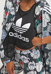 Person som bär en svart Adidas crop top och matchande jacka och leggings med blommönster i röd, vit, blå och grön detalj.