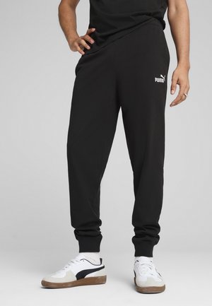 Personne portant un pantalon de survêtement Puma noir et des baskets blanches avec des rayures noires, debout avec une main sur la hanche sur fond gris.