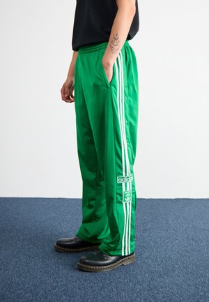 Person trägt grüne Adidas-Trainingshose mit weißen Streifen und schwarze Schuhe, steht auf blauem Teppich mit den Händen in den Taschen.
