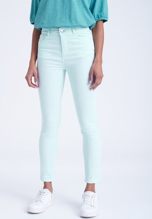 Person wearing light blue skinny jeans, teal short-sleeve top, and white sneakers, standing against a plain light background.