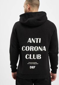 Czarna bluza z kapturem z kieszenią kangurką, z dużym białym napisem "ANTI CORONA CLUB" oraz dodatkowym tekstem poniżej. Materiał bawełniany.