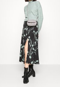 Žena v světlezeleném svetru, černé květinové midi sukni se rozparkem, s křížovou taškou a černými kotníkovými botami, stojící na bílém pozadí.