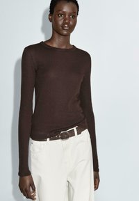 Maglione marrone a maniche lunghe, aderente, realizzato in tessuto a maglia, abbinato a pantaloni bianchi a vita alta e una cintura marrone scuro con fibbia argentata.