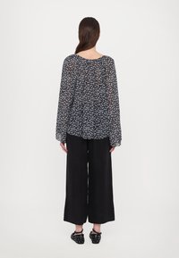 Femme aux longs cheveux bruns portant un chemisier noir à fleurs, un pantalon large noir et des chaussures noires cloutées, debout de dos.