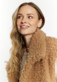 Tan, fluffy nepbont gilet met een brede kraag. Licht materiaal eronder, golvend haar gedeeltelijk zichtbaar. Zachte textuur en ontspannen ontwerp.