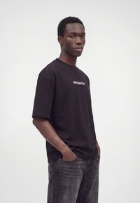 Jeune homme portant un t-shirt noir oversize avec texte et un jean noir délavé, debout les mains dans les poches devant un fond blanc.
