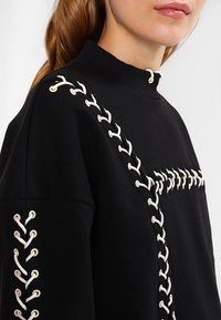 Musta collegepusero korkealla kauluksella, jossa valkoista nauhakuviota olkapäiltä ja edestä, valmistettu puuvillamateriaalista, jossa on teksturoidut yksityiskohdat.