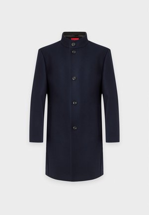 Homme portant une casquette noire, un manteau bleu marine foncé par-dessus un pull zippé noir, un pantalon noir à fines rayures et des chaussures en cuir noir, debout sur un fond blanc.