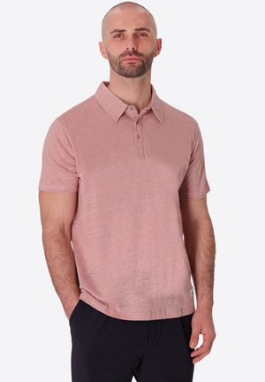 Uomo con barba corta che indossa una polo rosa chiaro a maniche corte e pantaloni neri, in piedi con una mano in tasca.