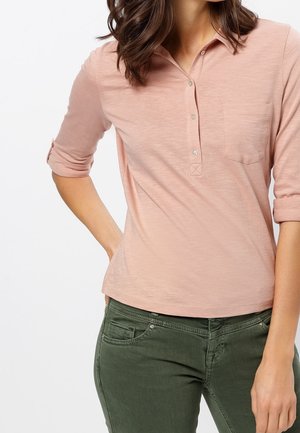 Kobieta ubrana w jasno różową koszulkę polo z długim rękawem z podwiniętymi rękawami i oliwkowo zielone spodnie, ręka oparta na biodrze.