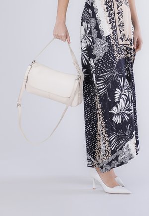 Kvinde iført et mønstret sort og beige maxiskørt med hvide højhælede sko, der holder en hvid læderhåndtaske i sin korte strop.