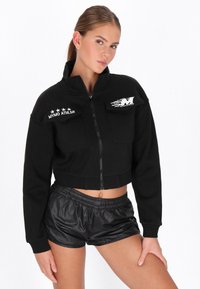 Geacă neagră scurtă cu fermoar, cu guler înalt, buzunare frontale și detalii cu stele albe. Asortată cu pantaloni scurți negri din material lucios.