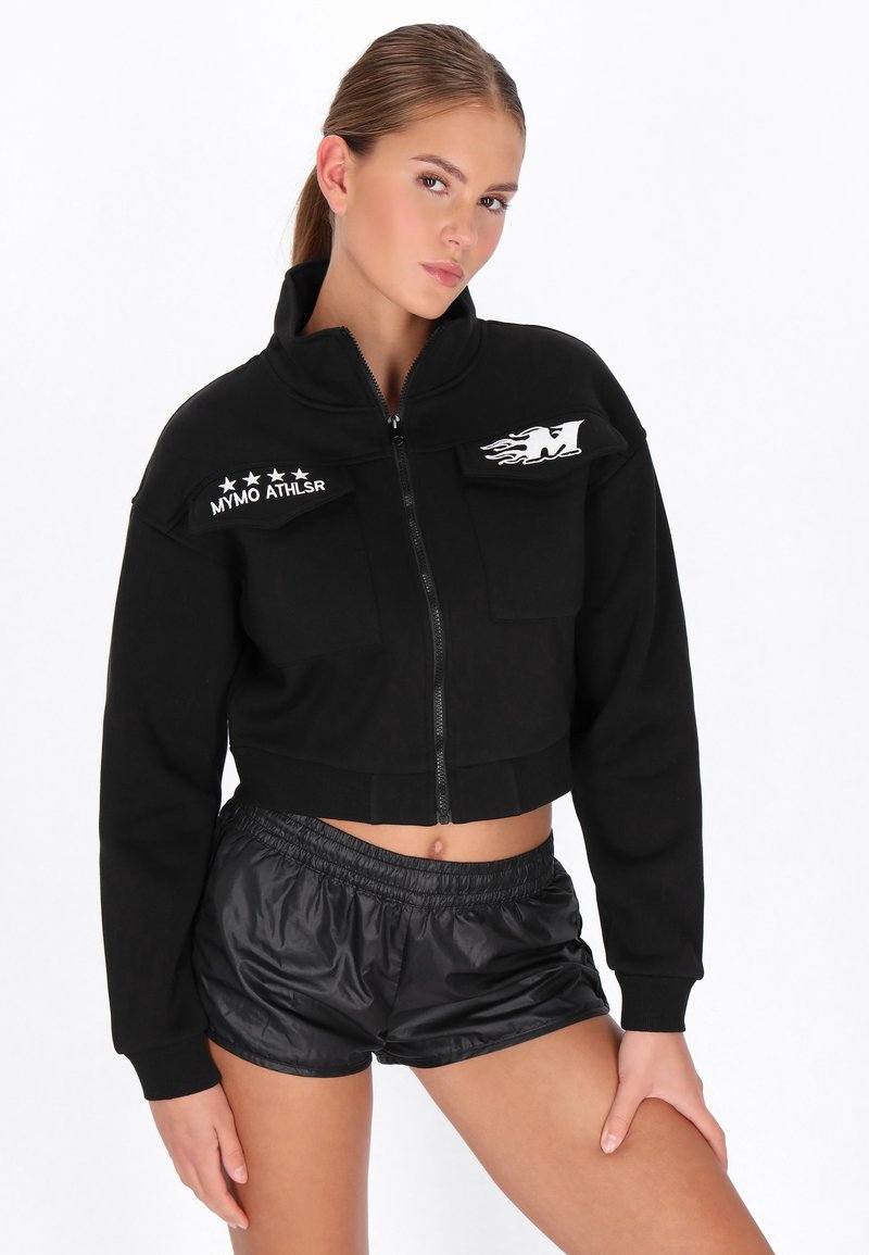 Chaqueta negra recortada con cremallera, cuello alto, bolsillos frontales y detalles de estrellas blancas. Combinada con pantalones cortos negros de material brillante.