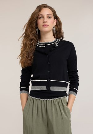 Young woman with long wavy hair wearing a black buttoned cardigan with white trim, green pleated skirt, and dangling earrings, hands in pockets.